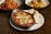 A wooden table displays a main dish of baked gnocchi with rich tomato sauce, melted cheese, and fresh herbs in a rustic metal dish, accompanied by slices of toasted bread on a white plate. In the background, two dishes filled with pasta and shrimp sit, featuring vibrant colors and garnished with greens.