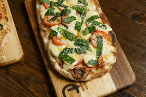 A freshly baked flatbread pizza on a wooden cutting board, topped with melted cheese, slices of ripe red tomatoes, and large pieces of fresh basil leaves, showcasing a golden-brown crust.