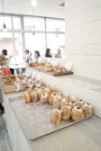 A bright, modern bakery with an assortment of pastries displayed on a wooden counter. Numerous small cream-filled pastries with decorative toppings rest on a metal tray in the foreground. Behind them are various bread and pastry items, including rolls and croissants. In the background, people are seated at tables near large windows, creating an inviting, social atmosphere. Overhead lights add to the cozy ambiance.