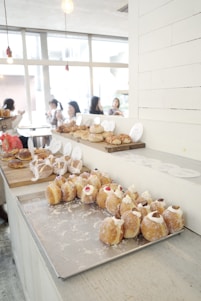 A bright, modern bakery with an assortment of pastries displayed on a wooden counter. Numerous small cream-filled pastries with decorative toppings rest on a metal tray in the foreground. Behind them are various bread and pastry items, including rolls and croissants. In the background, people are seated at tables near large windows, creating an inviting, social atmosphere. Overhead lights add to the cozy ambiance.