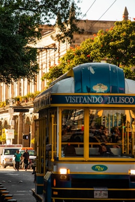 A vibrant street scene featuring a tour bus with 'Visitando Jalisco' written on it. The bus is blue and yellow with people visible inside. On the left, pedestrians and a white van are on the road. The background includes historical architecture with tall columns partially obscured by lush green trees and red flowers.
