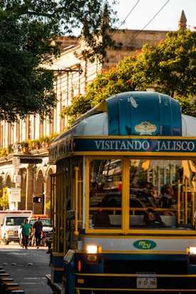 A vibrant street scene featuring a tour bus with 'Visitando Jalisco' written on it. The bus is blue and yellow with people visible inside. On the left, pedestrians and a white van are on the road. The background includes historical architecture with tall columns partially obscured by lush green trees and red flowers.