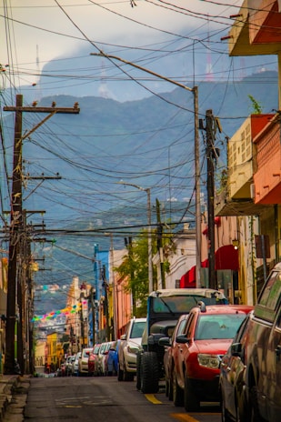 A bustling city street with colorful buildings and numerous cars parked along both sides. Overhead, numerous tangled electrical wires stretch between tall utility poles. In the distance, a misty mountain with communication towers can be seen under a cloudy sky.