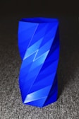 A vibrant blue, geometrically designed vase with angular facets and a twisted shape sits on a textured gray surface, creating a modern and artistic appearance.