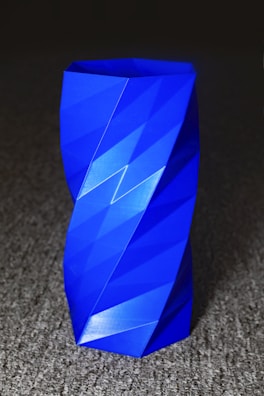 A vibrant blue, geometrically designed vase with angular facets and a twisted shape sits on a textured gray surface, creating a modern and artistic appearance.