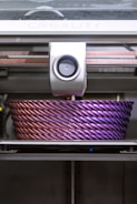A 3D printer is actively printing a large, intricately designed object with a rope-like texture. The object features a gradient of colors, transitioning from a metallic pink to a deep purple.