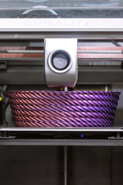 A 3D printer is actively printing a large, intricately designed object with a rope-like texture. The object features a gradient of colors, transitioning from a metallic pink to a deep purple.