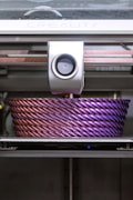 A 3D printer is actively printing a large, intricately designed object with a rope-like texture. The object features a gradient of colors, transitioning from a metallic pink to a deep purple.