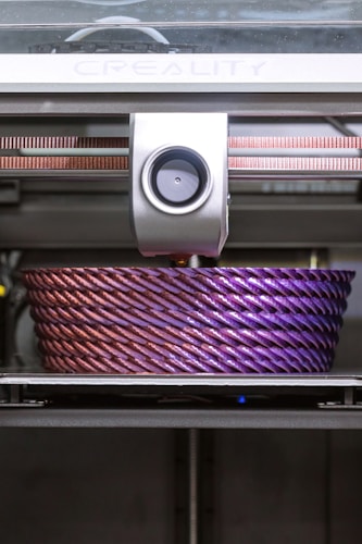 A 3D printer is actively printing a large, intricately designed object with a rope-like texture. The object features a gradient of colors, transitioning from a metallic pink to a deep purple.