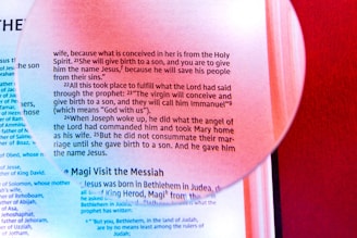 A magnifying glass overlays a page of religious text, highlighting a passage about the birth of Jesus. The text is printed in black on a light background with a distinct red hue from surrounding elements. The magnified section emphasizes certain words, creating a focus on that portion of the text.