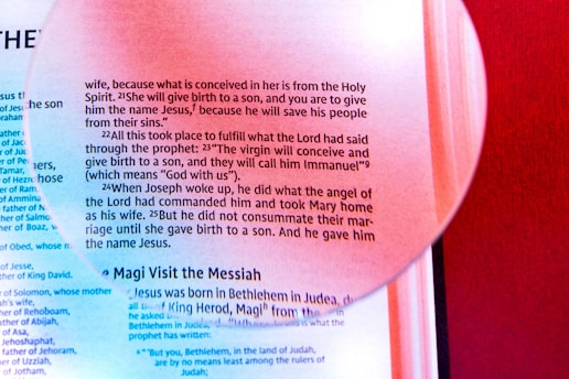 A magnifying glass overlays a page of religious text, highlighting a passage about the birth of Jesus. The text is printed in black on a light background with a distinct red hue from surrounding elements. The magnified section emphasizes certain words, creating a focus on that portion of the text.