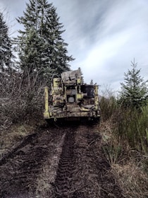 A large, rugged piece of machinery is positioned on a muddy forest trail, surrounded by lush green foliage and towering evergreen trees under a cloudy sky.