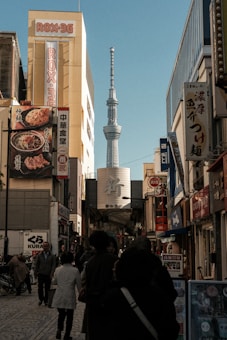 A bustling city street with numerous people walking along. Tall buildings with various advertisements and signs in Japanese line the sides of the street. In the background, a tall tower, possibly the Tokyo Skytree, rises prominently against a clear blue sky.
