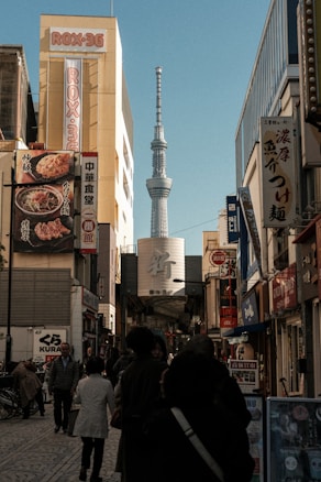 A bustling city street with numerous people walking along. Tall buildings with various advertisements and signs in Japanese line the sides of the street. In the background, a tall tower, possibly the Tokyo Skytree, rises prominently against a clear blue sky.