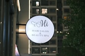 A round sign for a hair salon and beauty spa is mounted on the side of a building. The sign features elegant script text and a small heart design above the word 'Hi'. In the background, a building with illuminated windows is visible along with some trees on the right.