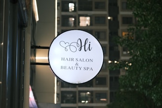 A round sign for a hair salon and beauty spa is mounted on the side of a building. The sign features elegant script text and a small heart design above the word 'Hi'. In the background, a building with illuminated windows is visible along with some trees on the right.