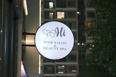 A round sign for a hair salon and beauty spa is mounted on the side of a building. The sign features elegant script text and a small heart design above the word 'Hi'. In the background, a building with illuminated windows is visible along with some trees on the right.