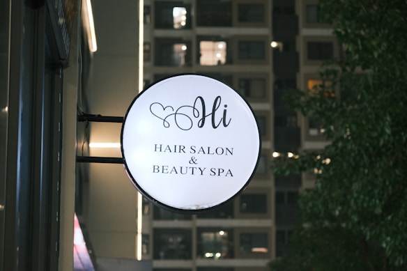 A round sign for a hair salon and beauty spa is mounted on the side of a building. The sign features elegant script text and a small heart design above the word 'Hi'. In the background, a building with illuminated windows is visible along with some trees on the right.