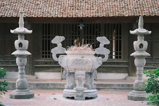 A traditional courtyard setting featuring a large ornate stone incense burner in the foreground flanked by two intricately carved stone lanterns. The background displays a classic wooden structure with a tiled roof and wooden window panels, evoking a sense of historical architecture.