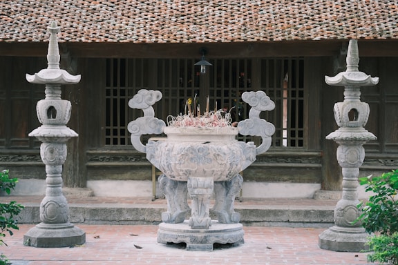 A traditional courtyard setting featuring a large ornate stone incense burner in the foreground flanked by two intricately carved stone lanterns. The background displays a classic wooden structure with a tiled roof and wooden window panels, evoking a sense of historical architecture.