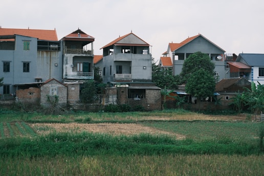 A cluster of residential buildings with grey and brown brick facades and red-orange roofs is situated adjacent to an overgrown field. The structures appear to be multi-storied with balconies and varying architectural designs. There are trees and greenery surrounding the buildings, contributing a rural feel to the setting.