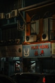 A nighttime street scene featuring shop signs and air conditioning units. The main sign is for 'TUCK HENG' with smaller text 'KEDAI UBAT'. There are other adjacent shop signs, including one partially visible sign saying 'WENG HOA'. The building is lit by external light sources, casting a warm glow on the windows and shopfronts. Several air conditioning units are visible mounted to the exterior walls.