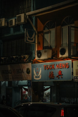 A nighttime street scene featuring shop signs and air conditioning units. The main sign is for 'TUCK HENG' with smaller text 'KEDAI UBAT'. There are other adjacent shop signs, including one partially visible sign saying 'WENG HOA'. The building is lit by external light sources, casting a warm glow on the windows and shopfronts. Several air conditioning units are visible mounted to the exterior walls.