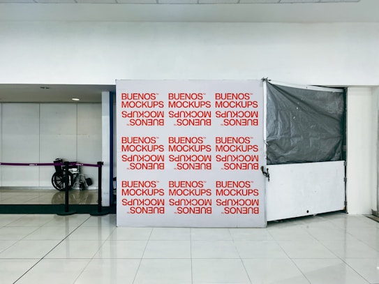 A tiled floor space with a large rectangular board printed with the words 'BUENOS MOCKUPS' repeatedly in red on a white background. Nearby is a purple rope barrier and a folded wheelchair. The wall and ceiling behind are plain and featureless, lending a sterile ambiance.