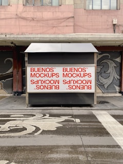 A street scene with a closed kiosk featuring large mockup advertisements in red on a white background. The kiosk is set against a backdrop of shuttered shopfronts and a building with a worn facade. The ground in front has artistic patterns and markings.