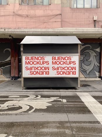 A street scene with a closed kiosk featuring large mockup advertisements in red on a white background. The kiosk is set against a backdrop of shuttered shopfronts and a building with a worn facade. The ground in front has artistic patterns and markings.