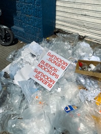 A pile of transparent plastic waste, paper, and a noticeable printed card with the text 'BUENOS MOCKUPS' lying on a sidewalk. Some cardboard and a closed metal shutter in the background next to a blue brick wall.