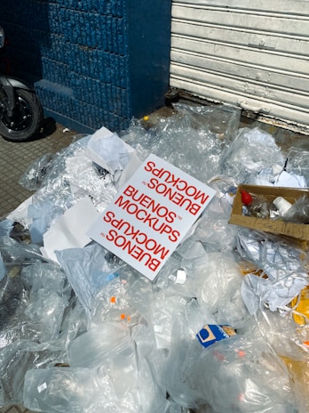 A pile of transparent plastic waste, paper, and a noticeable printed card with the text 'BUENOS MOCKUPS' lying on a sidewalk. Some cardboard and a closed metal shutter in the background next to a blue brick wall.