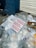 A pile of transparent plastic waste, paper, and a noticeable printed card with the text 'BUENOS MOCKUPS' lying on a sidewalk. Some cardboard and a closed metal shutter in the background next to a blue brick wall.