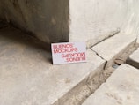 A printed card with the text 'BUENOS MOCKUPS' in bold orange letters is leaning against a rough, weathered concrete or stone wall. The surface beneath the card is uneven and cracked, resembling a construction or unfinished area. The overall setting appears industrial or urban.