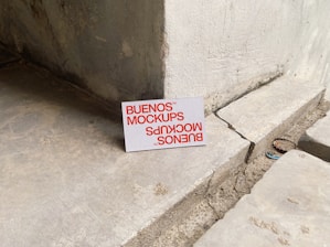 A printed card with the text 'BUENOS MOCKUPS' in bold orange letters is leaning against a rough, weathered concrete or stone wall. The surface beneath the card is uneven and cracked, resembling a construction or unfinished area. The overall setting appears industrial or urban.