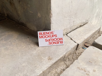 A printed card with the text 'BUENOS MOCKUPS' in bold orange letters is leaning against a rough, weathered concrete or stone wall. The surface beneath the card is uneven and cracked, resembling a construction or unfinished area. The overall setting appears industrial or urban.