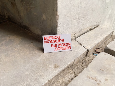 A printed card with the text 'BUENOS MOCKUPS' in bold orange letters is leaning against a rough, weathered concrete or stone wall. The surface beneath the card is uneven and cracked, resembling a construction or unfinished area. The overall setting appears industrial or urban.