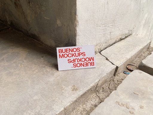 A printed card with the text 'BUENOS MOCKUPS' in bold orange letters is leaning against a rough, weathered concrete or stone wall. The surface beneath the card is uneven and cracked, resembling a construction or unfinished area. The overall setting appears industrial or urban.