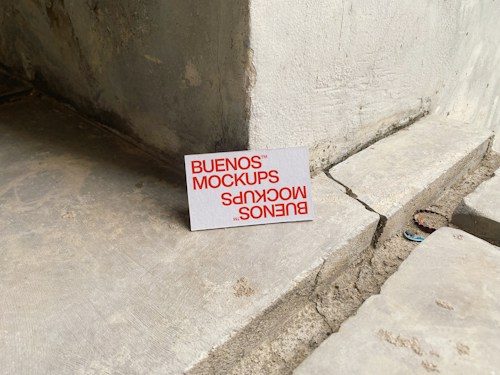 A printed card with the text 'BUENOS MOCKUPS' in bold orange letters is leaning against a rough, weathered concrete or stone wall. The surface beneath the card is uneven and cracked, resembling a construction or unfinished area. The overall setting appears industrial or urban.