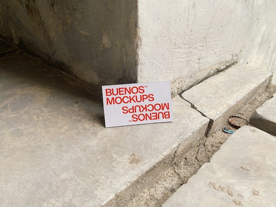 A printed card with the text 'BUENOS MOCKUPS' in bold orange letters is leaning against a rough, weathered concrete or stone wall. The surface beneath the card is uneven and cracked, resembling a construction or unfinished area. The overall setting appears industrial or urban.