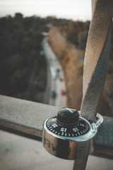 A combination lock is attached to a metal railing, with a blurred urban background showing a road lined with trees.