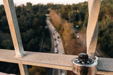 A combination lock is attached to a metal grate in the foreground. In the background, there is a road with cars driving along it, surrounded by trees with foliage in various shades of green and brown.