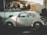 A vintage car is parked on the street with a 'For Sale' sign displayed on the window. The car, with its classic design, appears to be well-maintained and is parked in front of a house with a small garden and white wall. Sunlight creates soft shadows on the car and surroundings, adding a nostalgic feel.