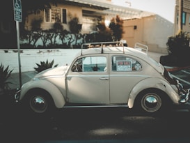 A vintage car is parked on the street with a 'For Sale' sign displayed on the window. The car, with its classic design, appears to be well-maintained and is parked in front of a house with a small garden and white wall. Sunlight creates soft shadows on the car and surroundings, adding a nostalgic feel.