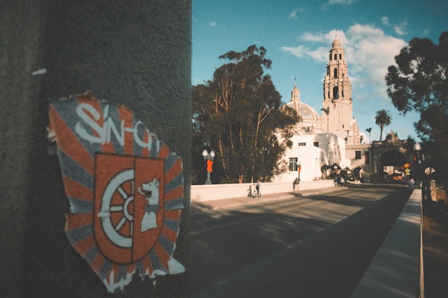 A sticker with a shield emblem and a stylized lion is affixed to a wall in the foreground. In the background, there is a grand historic building with a tall tower, surrounded by trees. A few people are walking and cycling on the road leading to the building under a blue sky with some clouds.