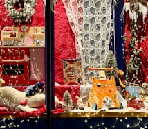 A festive display with toy kitchen sets, stuffed animals, and a variety of children's toys surrounded by decorative lights and a red backdrop. The scene is adorned with a snow-covered wreath and miniature Christmas trees, creating a cozy holiday atmosphere.