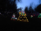 Colorful light displays in a park during dusk. LED lights create figures of elves and a colorful mound, possibly representing a pile of gifts or a festive scene. Trees and a darkening sky form the backdrop.