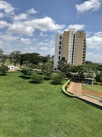 A tall apartment building with the name 'Mi Vida' is situated next to a well-maintained green lawn and trees. The sky is bright blue with scattered white clouds, creating a serene atmosphere. A pathway bordering the lawn leads to the building entrance.