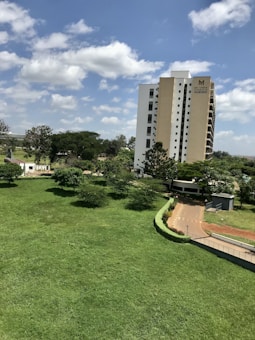 A tall apartment building with the name 'Mi Vida' is situated next to a well-maintained green lawn and trees. The sky is bright blue with scattered white clouds, creating a serene atmosphere. A pathway bordering the lawn leads to the building entrance.