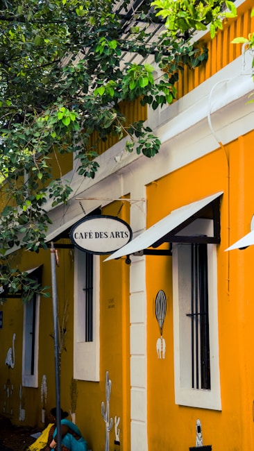 A quaint cafe with a bright yellow facade features a sign reading 'Caf&eacute; des Arts'. The building is adorned with black iron bars on the white-framed windows and artistic wall paintings depicting plants and abstract designs. Lush green leaves from a nearby tree partly shade the exterior, adding a natural touch to the urban setting.